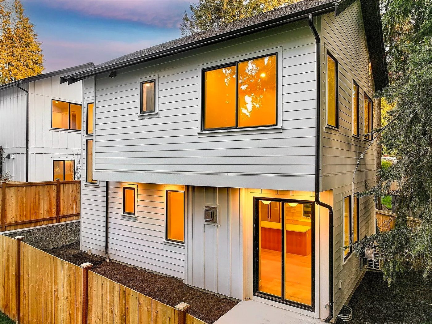 A two-level backyard home with white lap siding, lit up at dusk